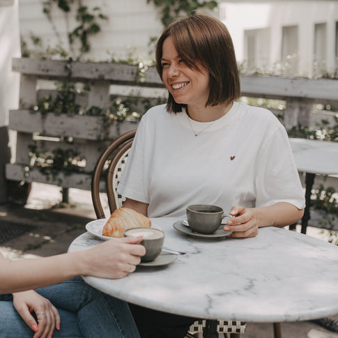 Frau im weißen T-Shirt mit Herz sitzt im Café mit Kaffee und Croissant