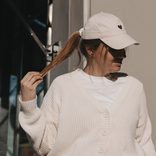Beige Cap mit schwarzem Herz-Stick, getragen von Frau im Sonnenlicht vor Eulenschnitt Store