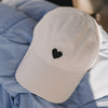 Beige Cap mit schwarzem Herz-Stick liegend auf blauer Jacke