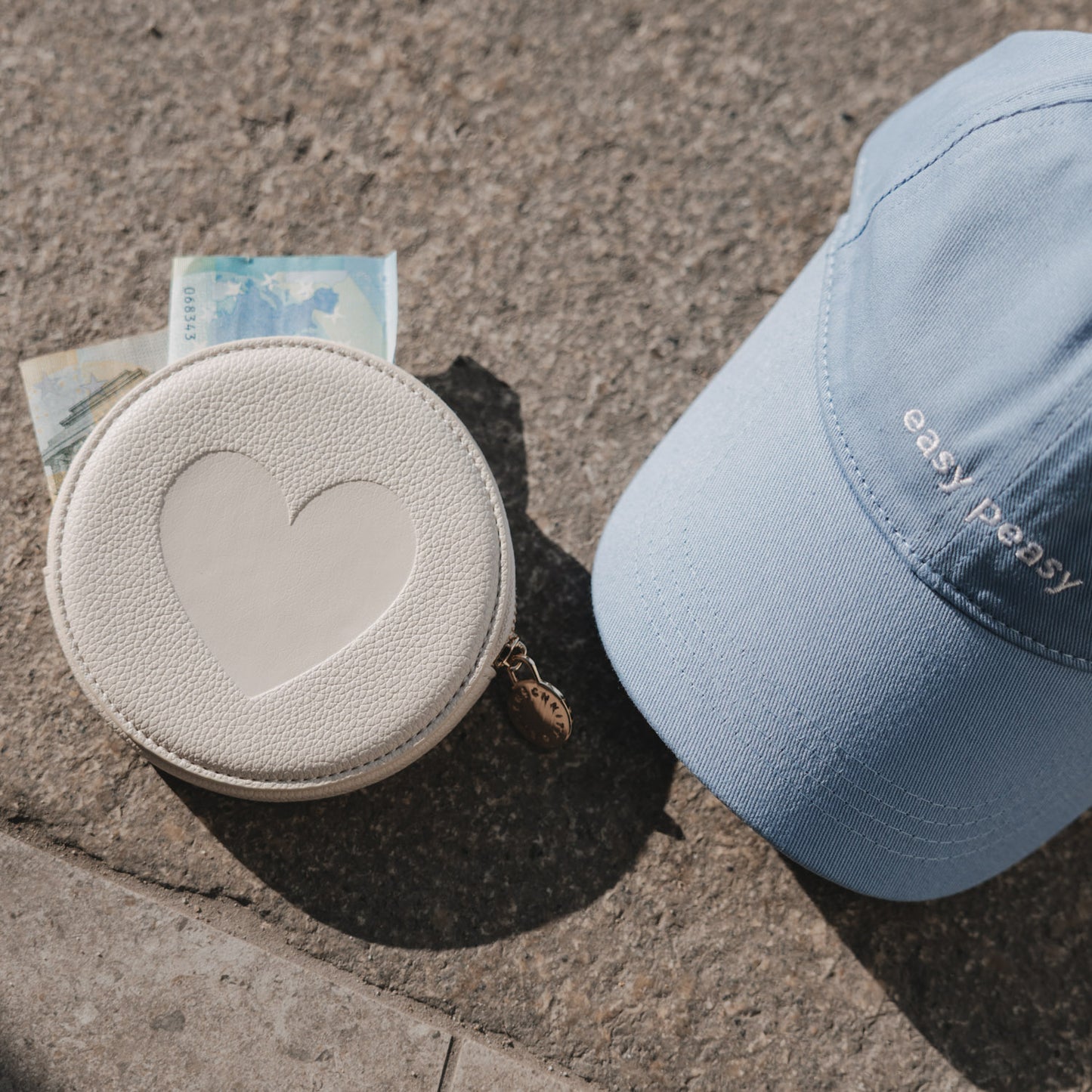 Hellblaue Cap „easy peasy“ neben rundem Portemonnaie mit Herzmotiv auf Steinboden