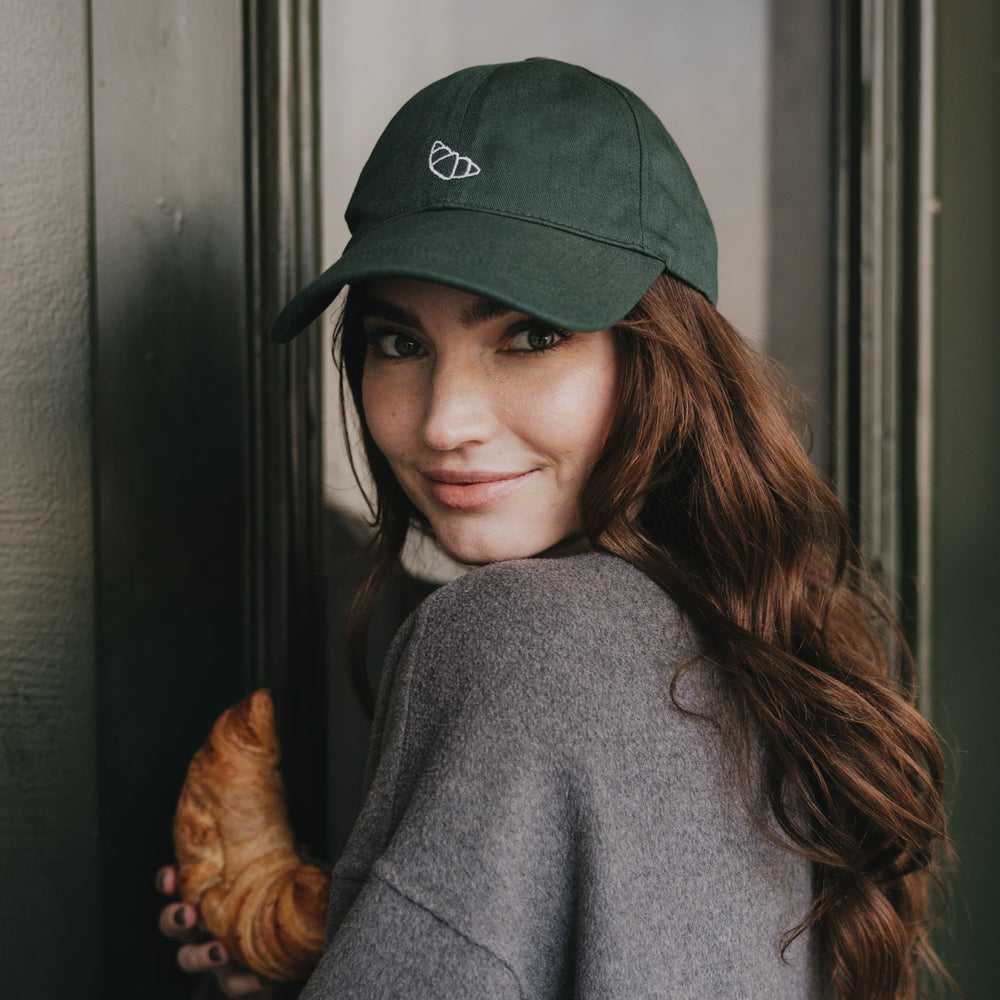Frau trägt dunkelgrüne Cap mit Croissant-Stickerei und hält Croissant in der Hand