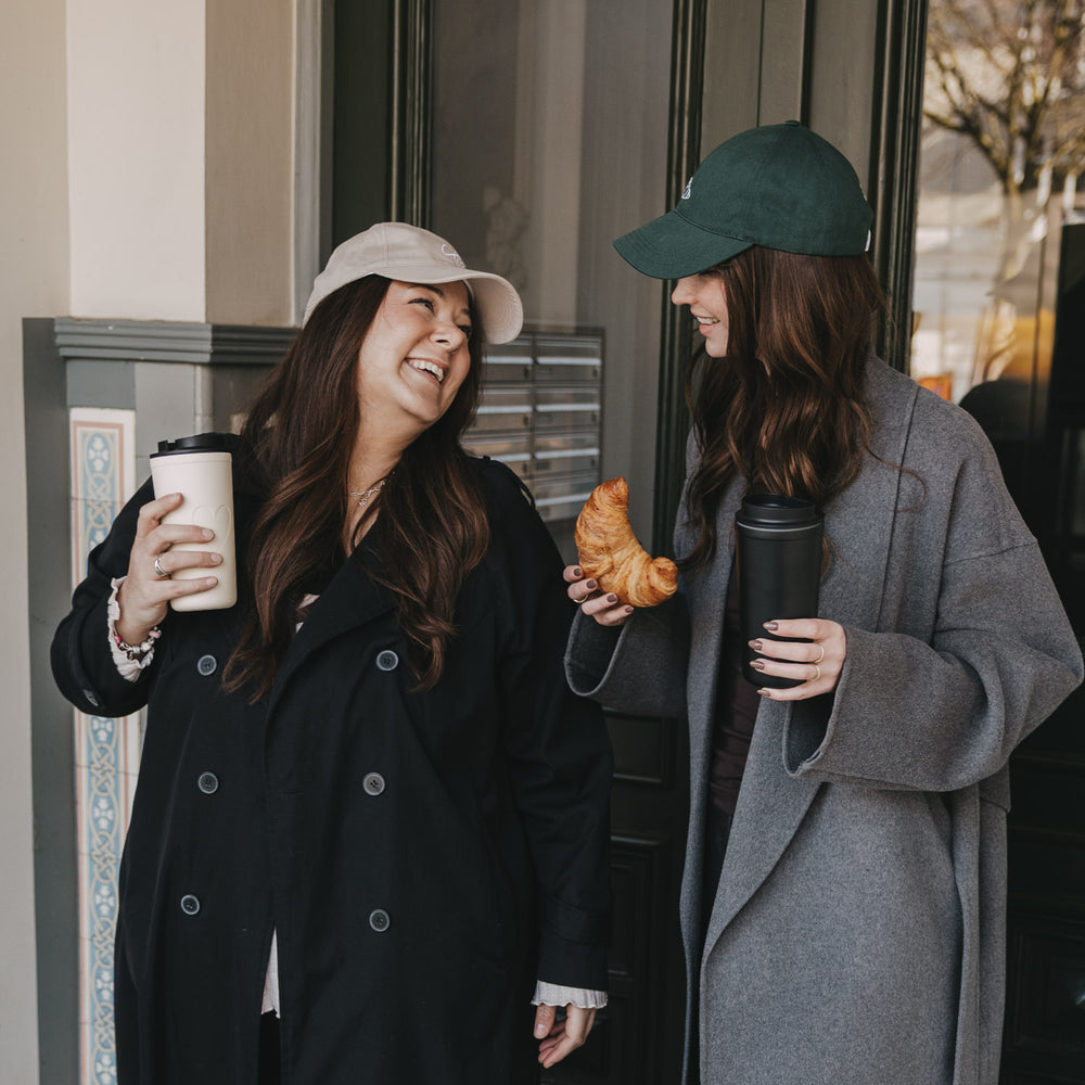 Zwei Frauen mit Caps und Coffee-to-go, eine trägt dunkelgrüne „Breakfast Club“ Cap