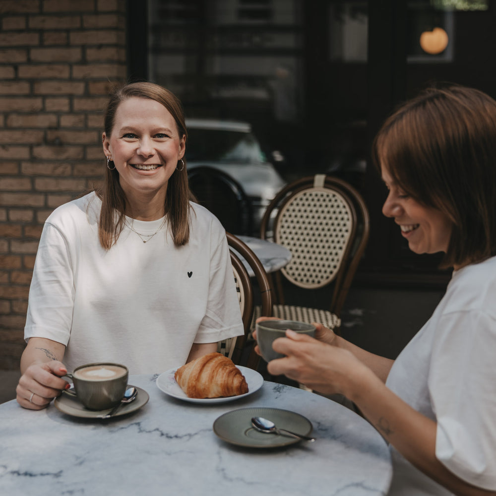 Zwei Frauen im Café mit Kaffee, eine trägt weißes T-Shirt mit kleinem Herz-Print