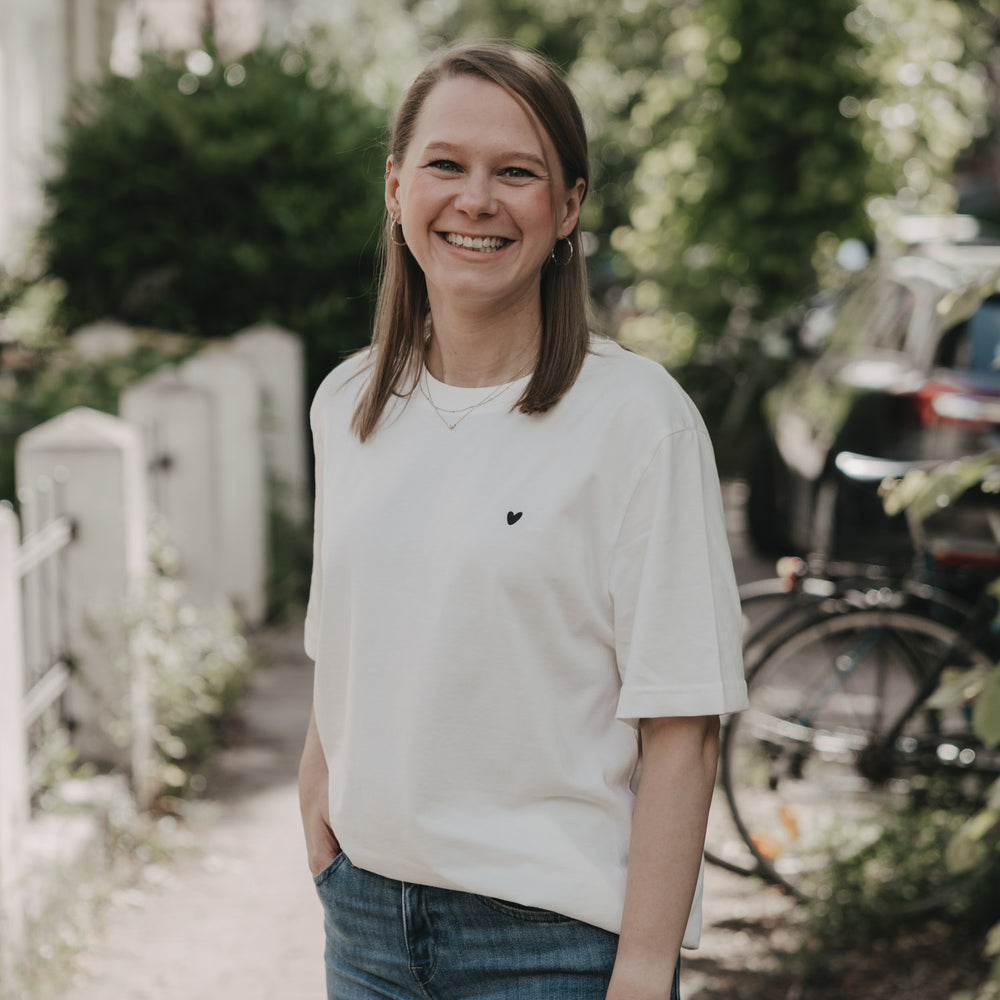 Frau im weißen T-Shirt mit Herz-Print steht lächelnd im Grünen
