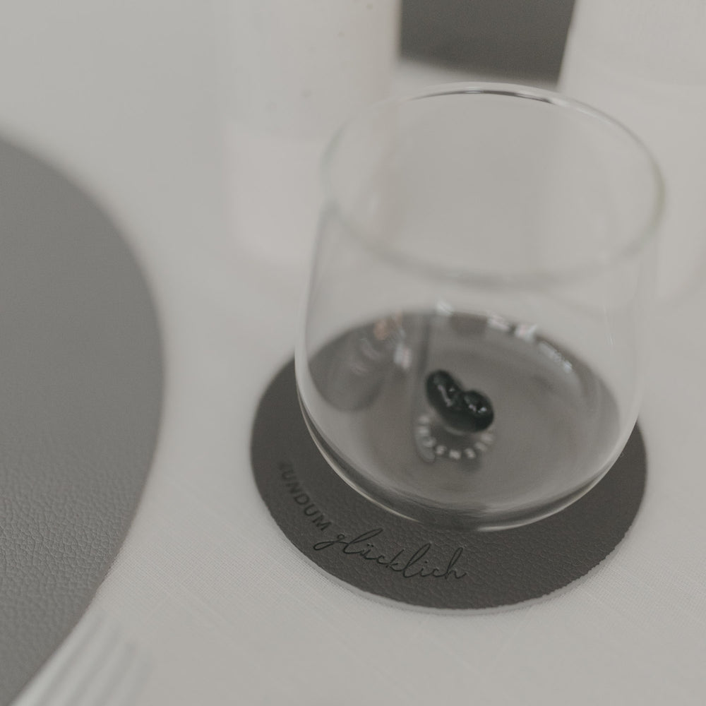 Grauer Untersetzer „Rundum glücklich“ mit Glas darauf, modernes Design auf hell gedecktem Tisch.