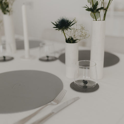 Grauer Untersetzer „Rundum glücklich“ auf elegant gedecktem Tisch mit weißen Vasen und grauen Platzsets.