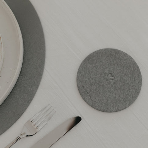 Dunkelgrauer Herz-Untersetzer neben Besteck und grauem Platzset auf weißer Tischdecke.