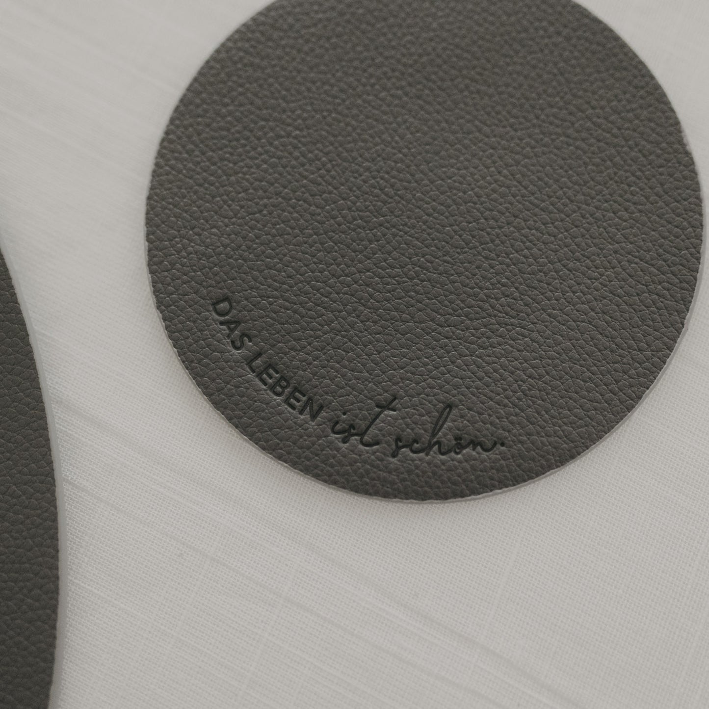 Runder Untersetzer in Dunkelgrau mit Prägung ‚Das Leben ist schön‘ auf weißer Tischdecke.