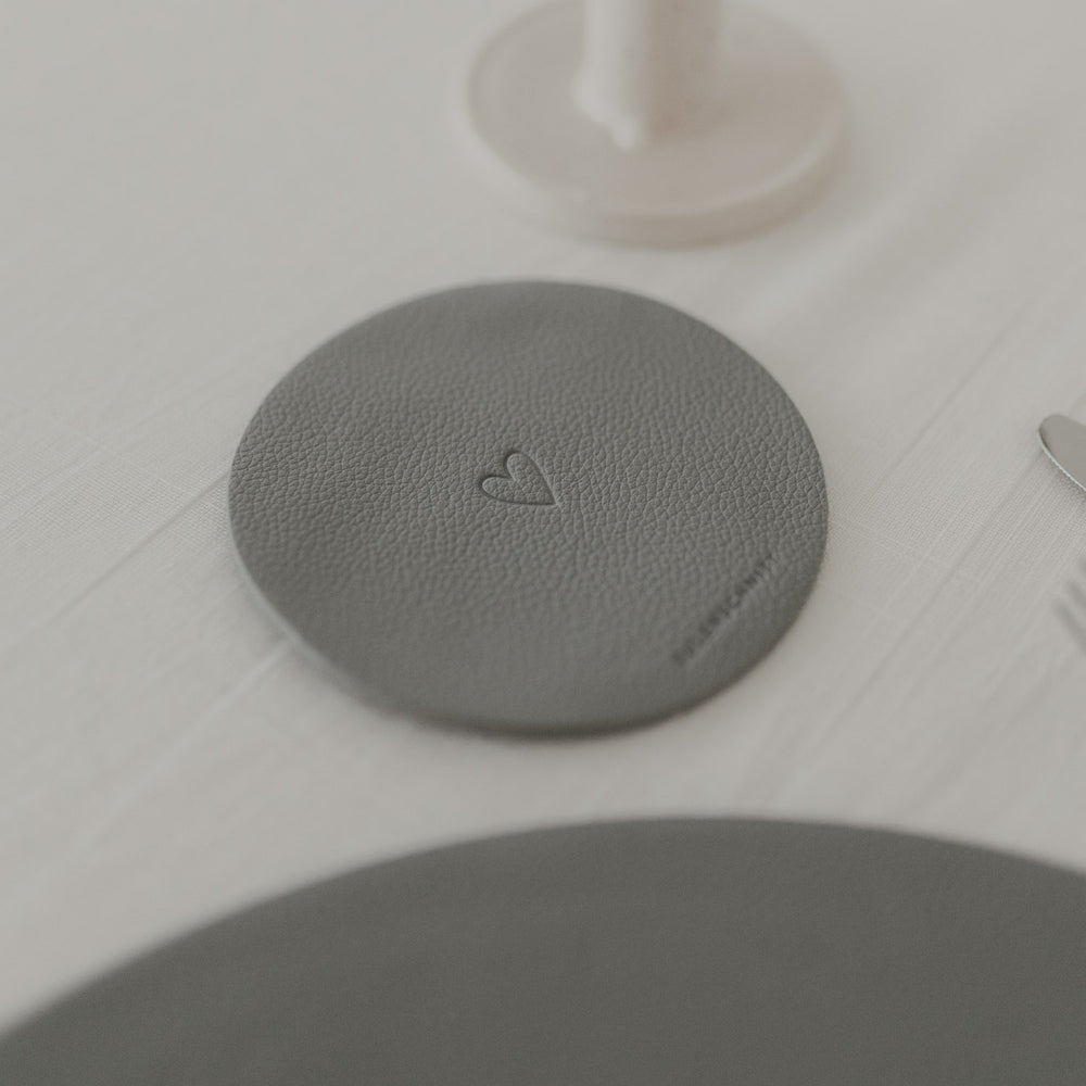 Runder hellgrauer Untersetzer mit geprägtem Herzmotiv, platziert auf weißer Tischdecke.