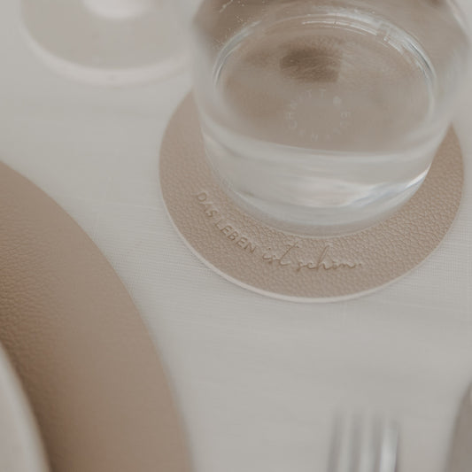 Runder Untersetzer in Beige mit Prägung „Das Leben ist schön“ unter einem Wasserglas auf weißem Tisch.