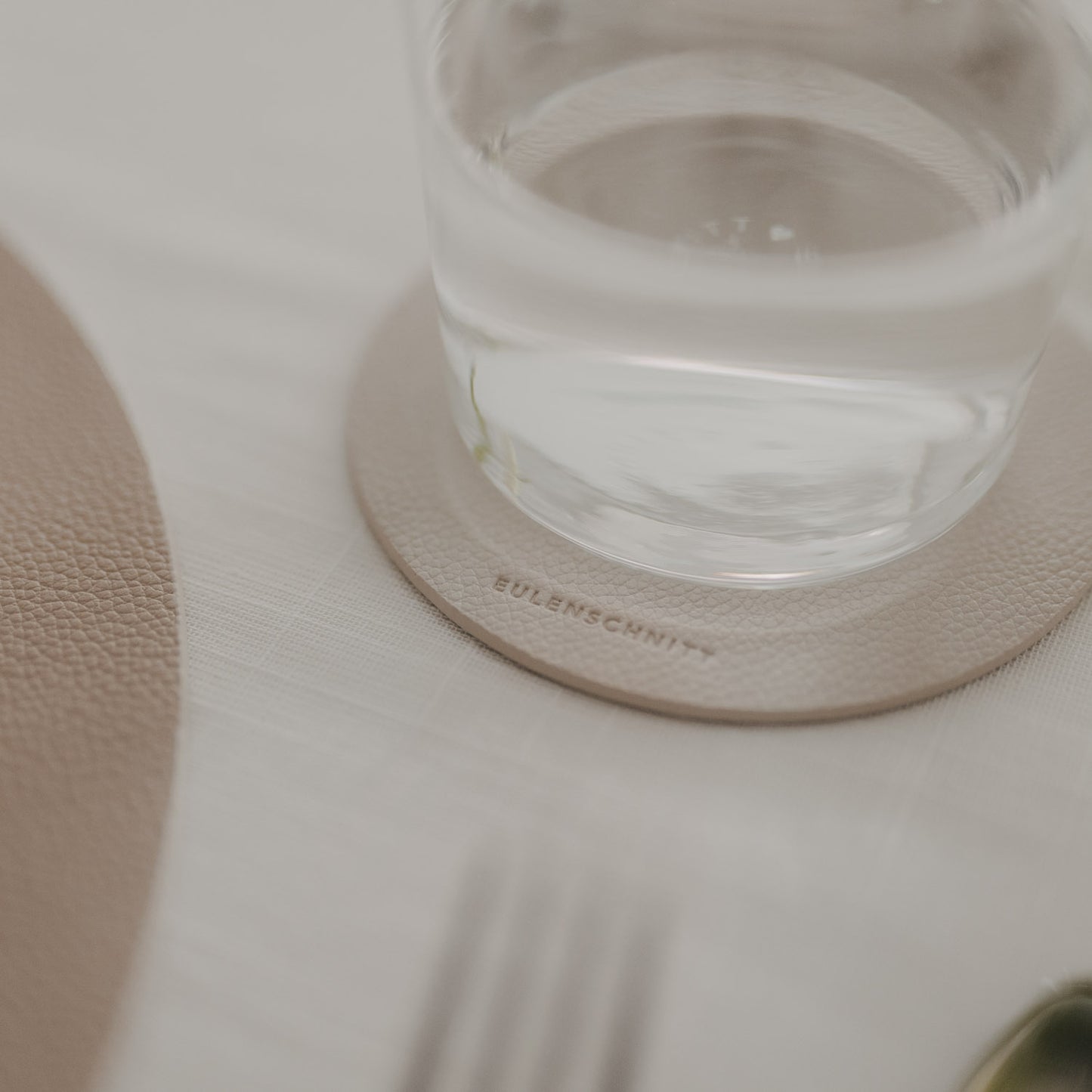 Wasserglas auf beigem Untersetzer mit Eulenschnitt-Prägung auf hell gedecktem Tisch.