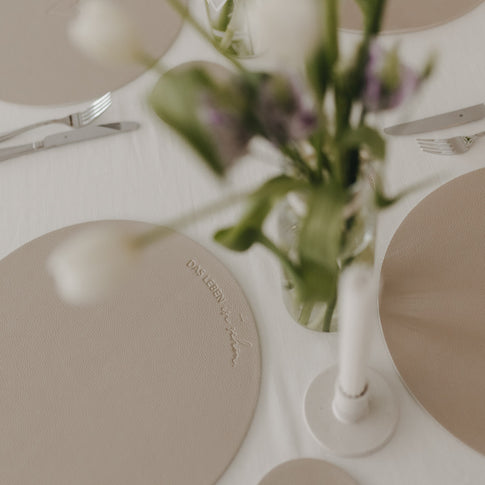 Elegante Tischdeko mit beigen Platzsets aus Kunstleder und Blumenvase auf weißer Tischdecke.