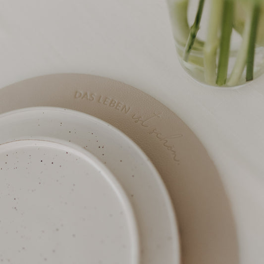 Beiges Platzset mit Prägung ‚Das Leben ist schön‘ unter weißem Keramikteller auf gedecktem Tisch.