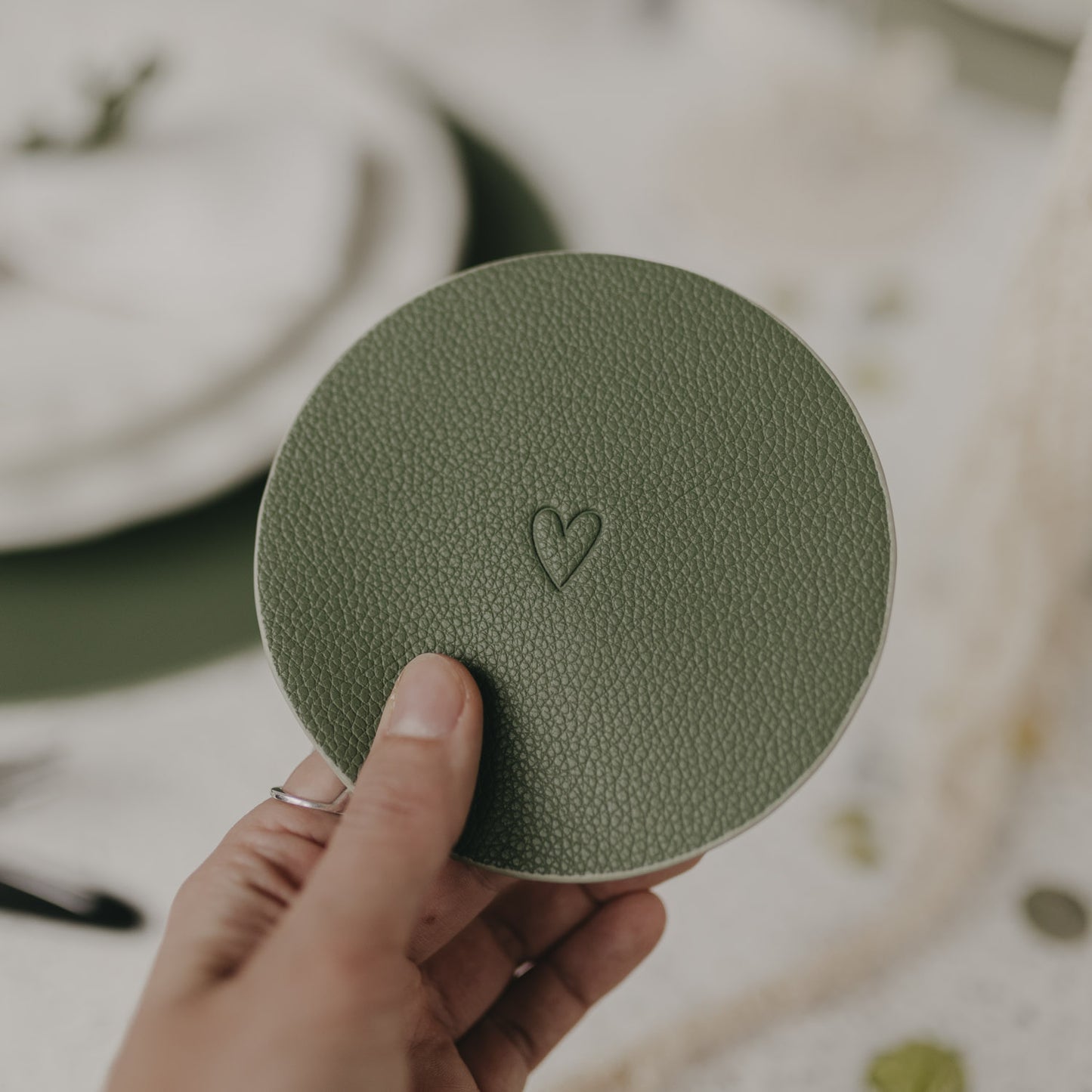 Hand hält grünen Untersetzer mit geprägtem Herzmotiv über gedecktem Tisch.