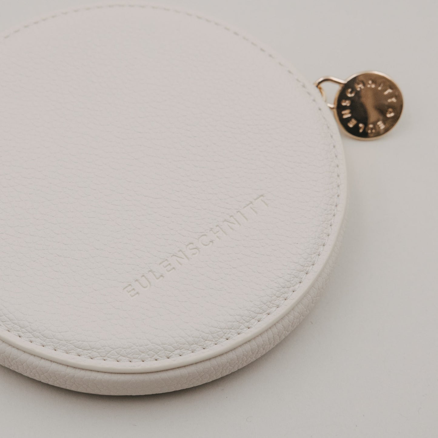 Rundes Portemonnaie in Creme mit geprägtem Eulenschnitt Logo und goldenem Reißverschlussanhänger.