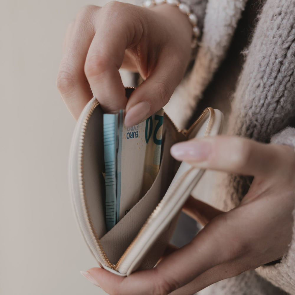 Geöffnetes rundes Portemonnaie in Creme mit Geldscheinen im Innenfach und goldenem Reißverschluss.