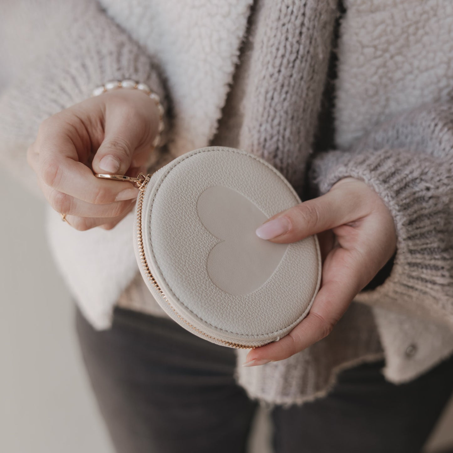 Rundes cremefarbenes Portemonnaie mit Herzmotiv, in Händen gehalten mit goldenem Reißverschluss.