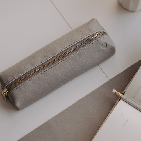 Graue Federtasche mit Herzprägung auf Schreibtisch neben Notizbuch und Bleistift