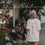Frau im weißen „Coffee Lover“-T-Shirt und rosa Cap mit Iced Coffee vor einem Blumenladen.