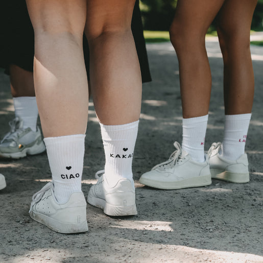 Mehrere Personen stehen gemeinsam auf einem Kiesweg. Sie tragen weiße Sneaker und verschiedene weiße Socken mit schwarzen und bunten Designs an den Fersen. Im Vordergrund ist ein Paar Socken mit der Aufschrift 'CIAO' und 'KAKAO' sowie kleinen schwarzen Herzen darüber gut zu erkennen.