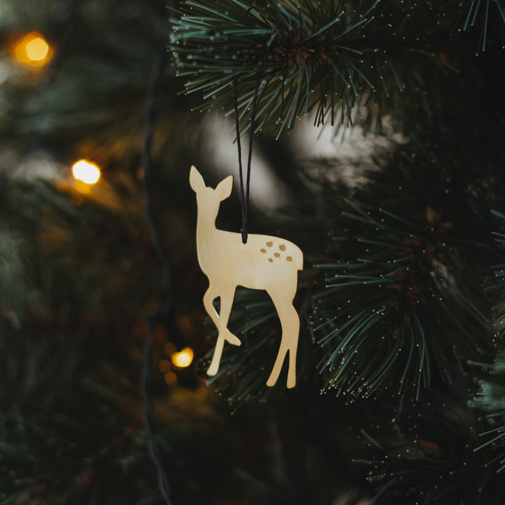 Goldener Rehanhänger  mit schwarzem Band an grünem Tannenzweig, umgeben von warmem Lichterglanz.