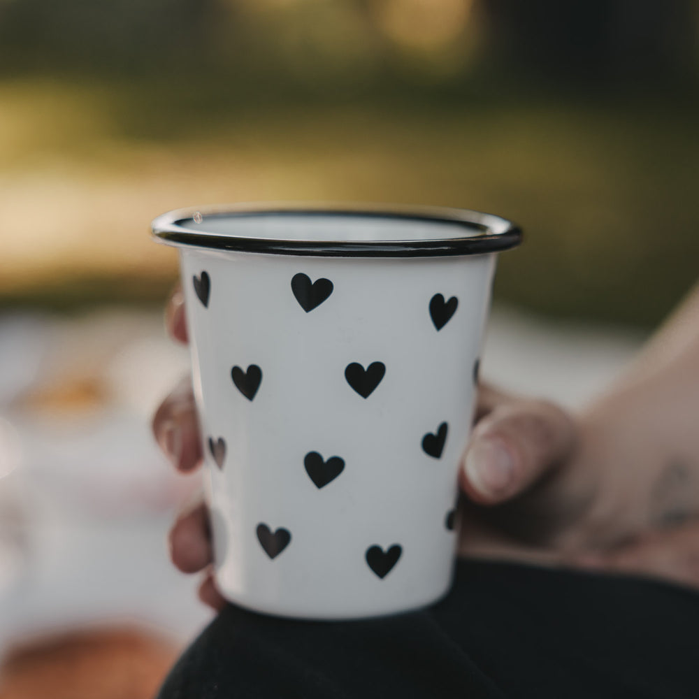 Weißer Emaille-Becher mit schwarzem Herzmuster wird in der Hand gehalten.
