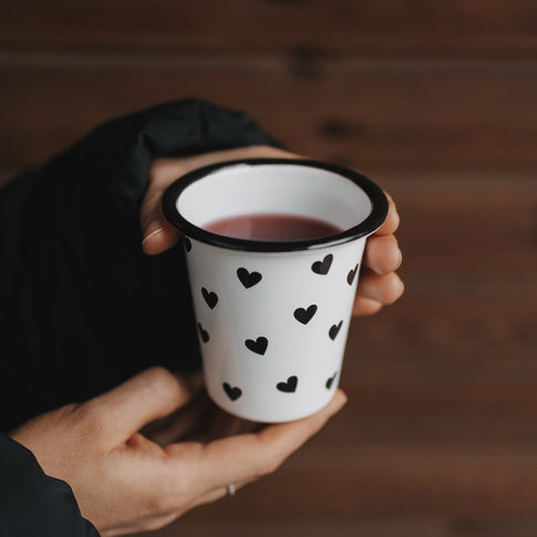 Emaillebecher mit schwarzen Herzen wird in beiden Händen gehalten, gefüllt mit warmem Getränk.