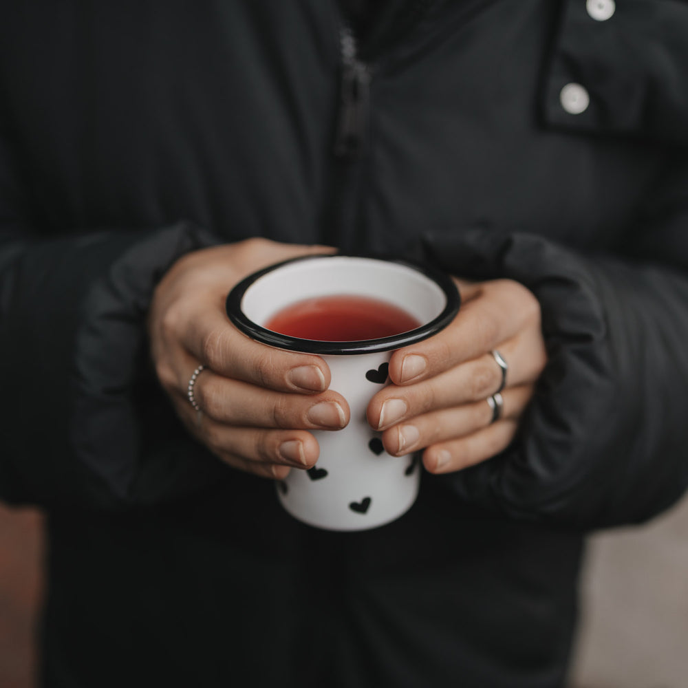 Person hält Emaillebecher mit schwarzen Herzen und warmem Getränk in beiden Händen vor dunkler Jacke.