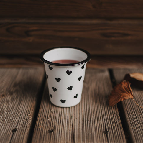 Weißer Emaillebecher mit schwarzen Herzen steht auf Holzuntergrund, gefüllt mit rotem Getränk.