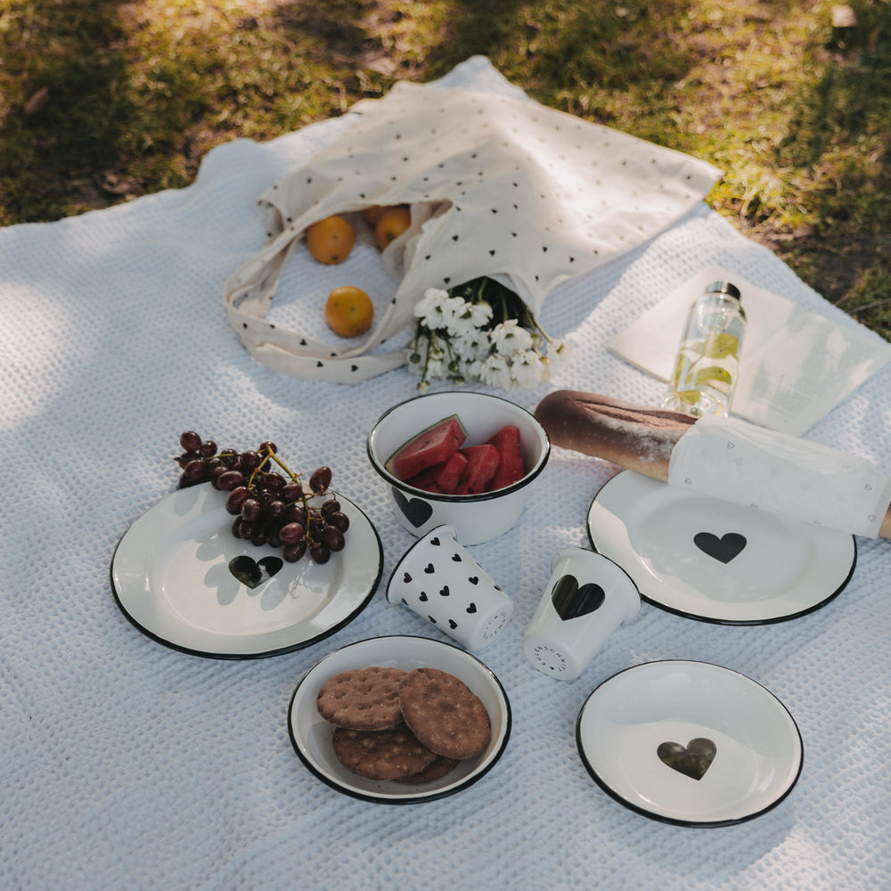 Weißer Emaille-Becher mit großem schwarzem Herz im sommerlichen Picknick-Setting mit passendem Emaille-Geschirr.