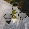 Weißer Emaille-Becher mit großem schwarzem Herz neben Trinkflasche und Herzbecher auf einer Picknickdecke.