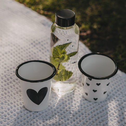 Weißer Emaille-Becher mit großem schwarzem Herz neben Trinkflasche und Herzbecher auf einer Picknickdecke.