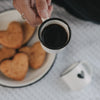 Weiße Emaille-Espressotasse mit schwarzem Herz, gefüllt mit Espresso, in der Hand gehalten, mit herzförmigen Keksen im Hintergrund.