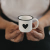 Weiße Emaille-Espressotasse mit schwarzem Herz wird mit beiden Händen vor neutralem Hintergrund gehalten.