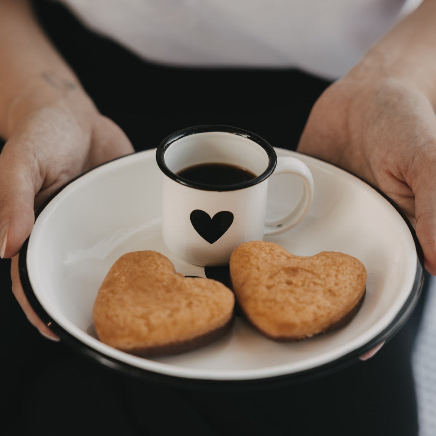 Weiße Emaille-Espressotasse mit schwarzem Herz und Espresso auf einem Emaille-Teller mit zwei herzförmigen Keksen.