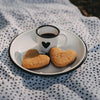 Weiße Emaille-Espressotasse mit schwarzem Herz und Espresso auf einem Emaille-Teller im sommerlichen Picknick-Setting mit herzförmigen Keksen.