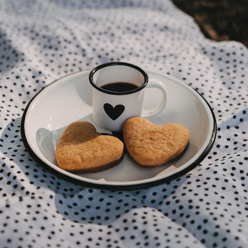 Weiße Emaille-Espressotasse mit schwarzem Herz und Espresso auf einem Emaille-Teller im sommerlichen Picknick-Setting mit herzförmigen Keksen.