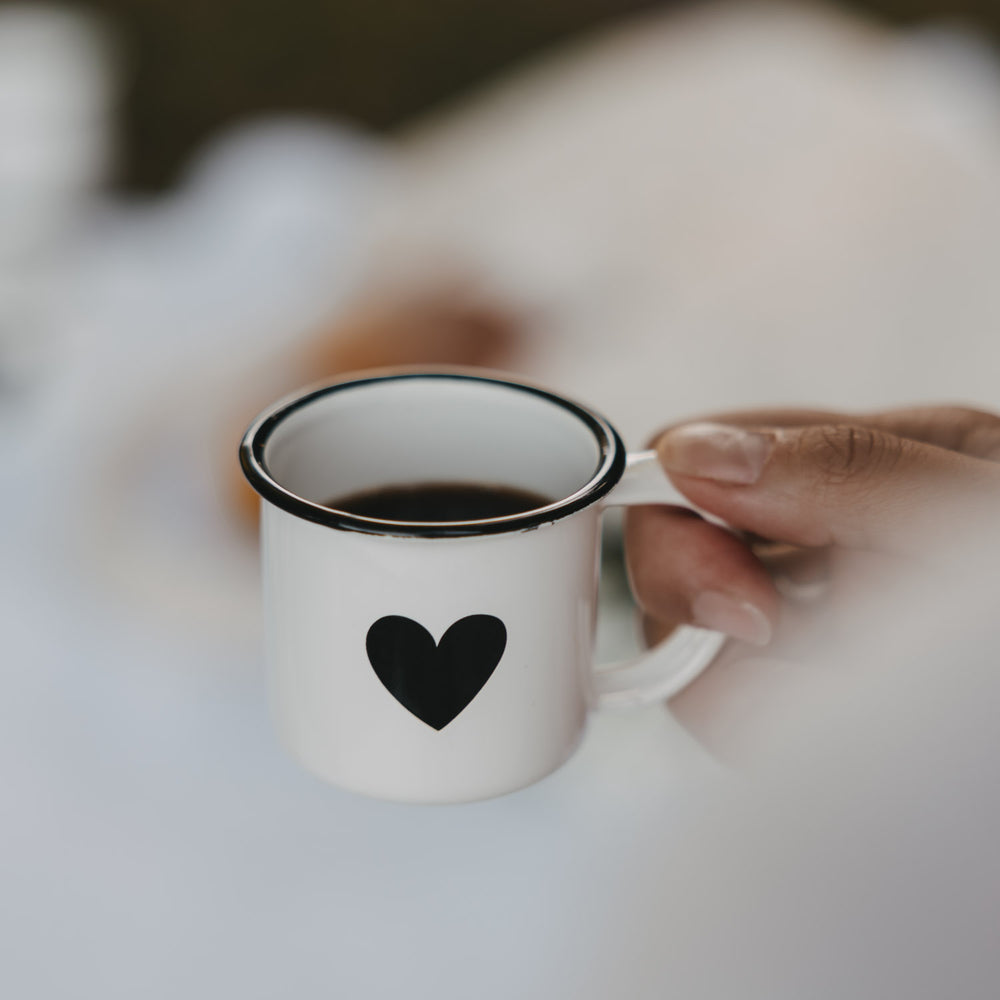 Weiße Emaille-Espressotasse mit schwarzem Herz, gefüllt mit Espresso, wird am Henkel gehalten.