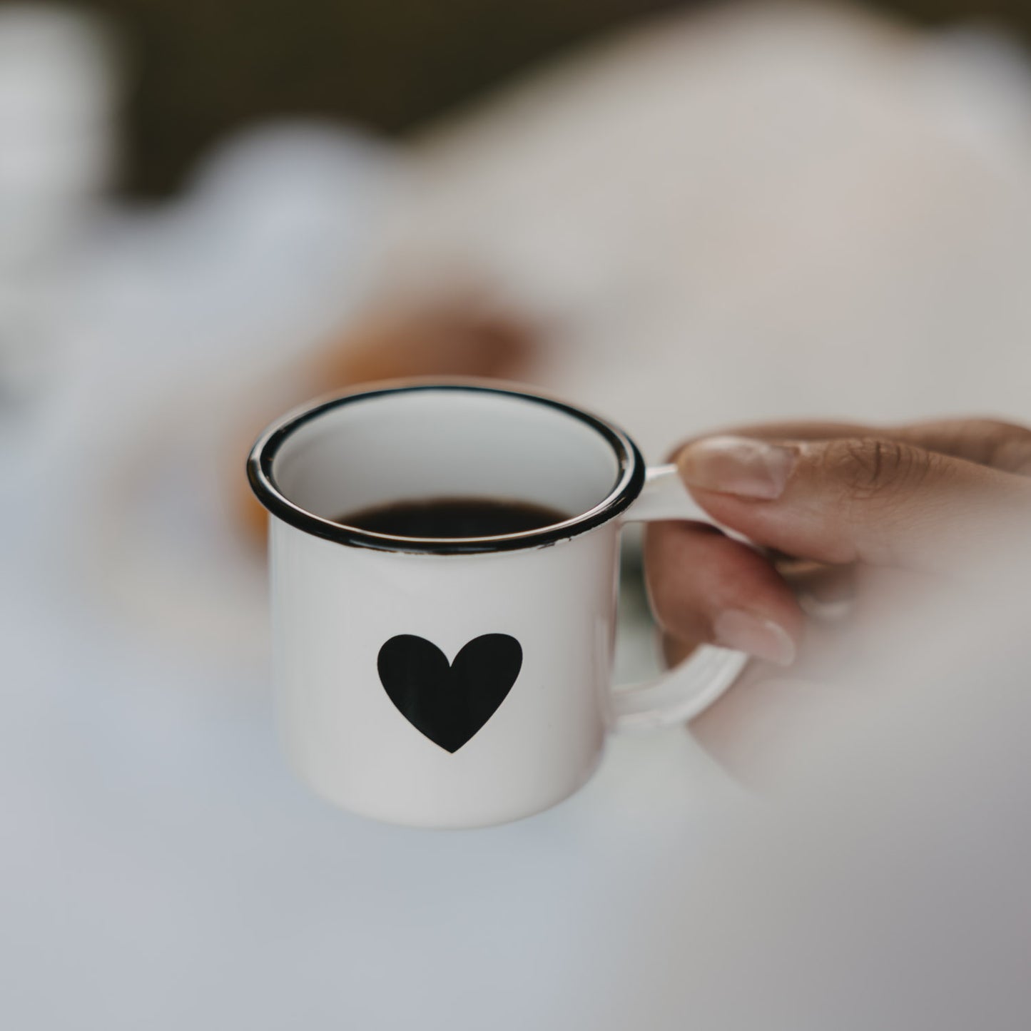 Weiße Emaille-Espressotasse mit schwarzem Herz, gefüllt mit Espresso, wird am Henkel gehalten.
