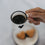 Weiße Emaille-Espressotasse mit schwarzem Herz, gefüllt mit Espresso, über einem Teller mit herzförmigen Keksen gehalten.