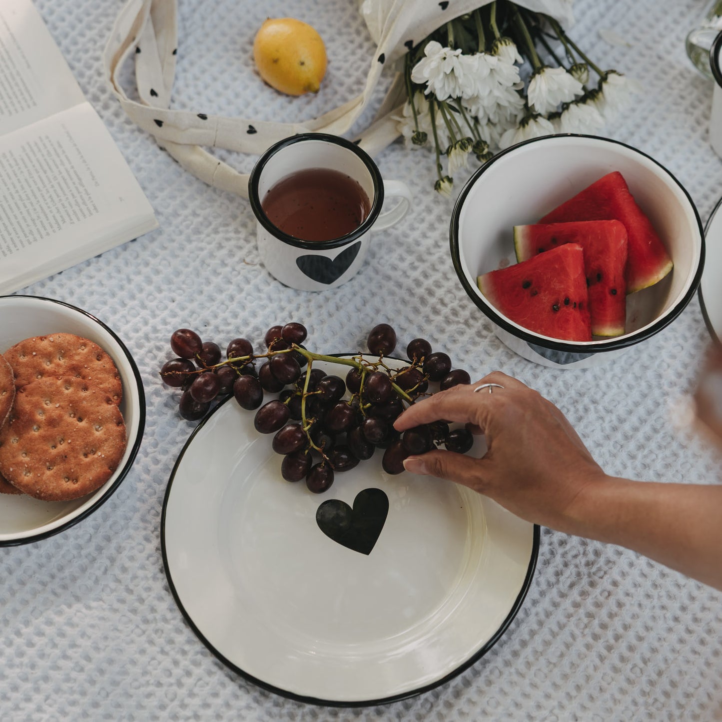 Weißer Emaille-Speiseteller mit schwarzem Herz im sommerlichen Picknick-Setting mit Weintrauben, Wassermelone und Emaille-Geschirr.