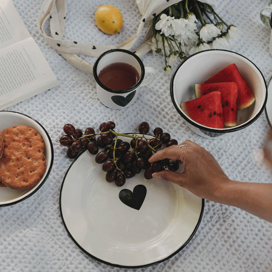 GIF zeigt den weißen Emaille-Speiseteller mit schwarzem Herz im sommerlichen Picknick-Setting mit Weintrauben und weiterem Emaille-Geschirr.