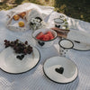 Emaille-Geschirr mit schwarzem Herz im sommerlichen Picknick-Setting auf einer hellen Decke.