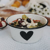 Weiße Emaille-Schale mit schwarzem Herz, gefüllt mit Salat, Feta und Oliven.