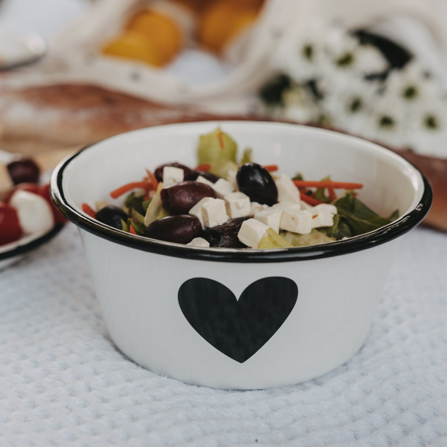 Weiße Emaille-Schale mit schwarzem Herz, gefüllt mit Salat, Feta und Oliven.