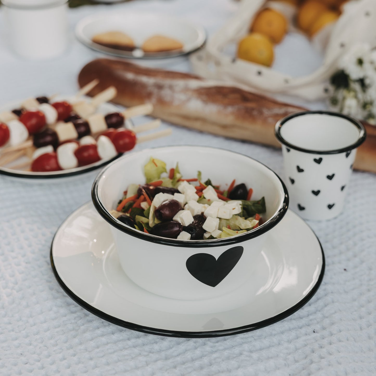Weiße Emaille-Schale mit schwarzem Herz auf passendem Emaille-Teller, gefüllt mit Salat, im sommerlichen Picknick-Setting.