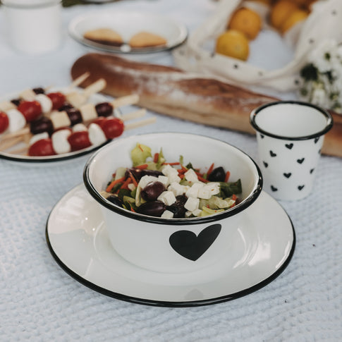 Weiße Emaille-Schale mit schwarzem Herz auf passendem Emaille-Teller, gefüllt mit Salat, im sommerlichen Picknick-Setting.