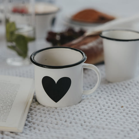 Weiße Emaille-Tasse mit schwarzem Herz auf einer hellen Decke, neben einem Buch und Frühstückszubehör.