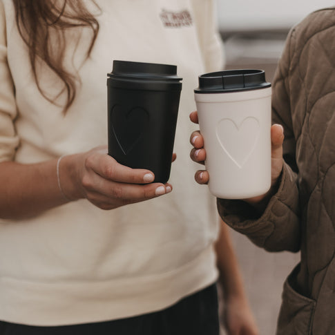 Schwarzer und weißer To-Go-Becher mit Herzmotiv und Deckel von Eulenschnitt in den Händen zweier Frauen