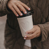 Cremefarbener To-Go-Becher mit Herzmotiv und schwarzem Deckel wird von einer Frau verschlossen